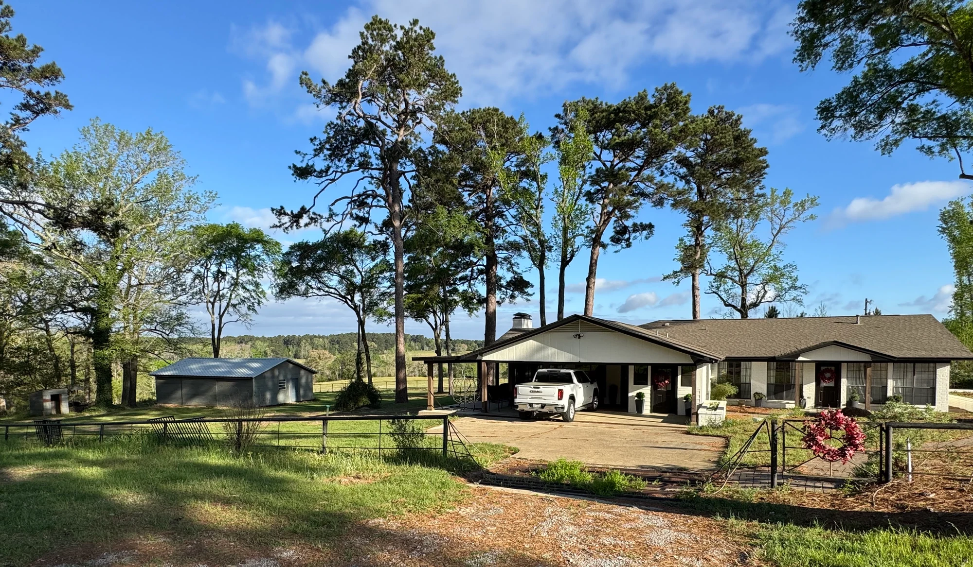 The single-story retreat house behind a fence, with tall trees and a covered parking area